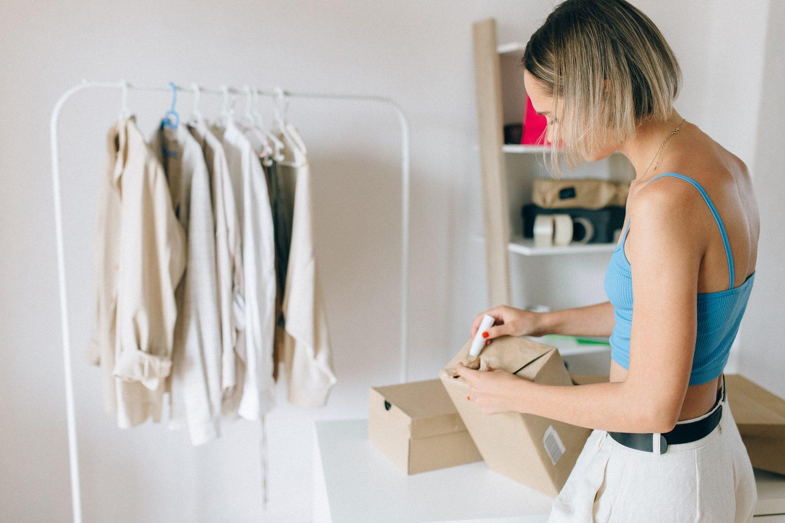 Instagram seller packing a clothing order into a courier box beside a garment rail